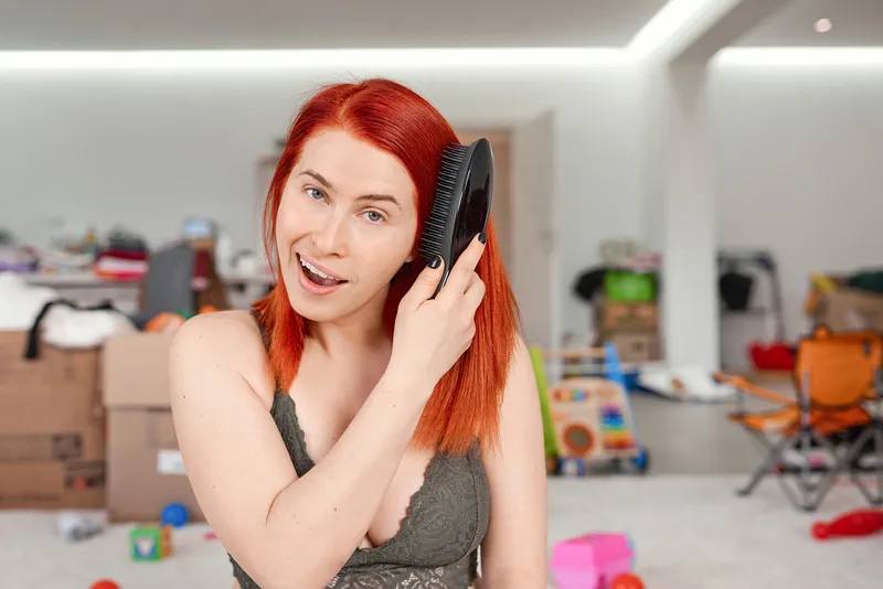 Smiling red-haired woman brushing her hair in a cluttered room.